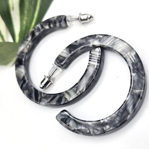 May include: A pair of C-shaped hoop earrings featuring a marbled pattern in shades of black, gray, and white. The earrings have a smooth, polished finish and include silver-toned metal posts with clear plastic earring backs for secure closure.