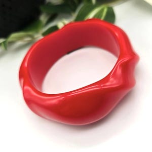 May include: A bright red bangle bracelet with a distinctive, undulating design. The glossy, solid-coloured bracelet is a striking accessory. The bracelet is a fashionable item.