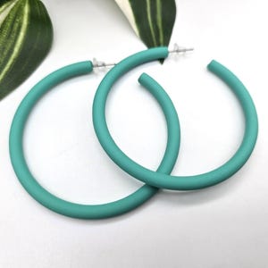 Op de afbeelding: Een paar turquoise oorringen. De oorbellen hebben een effen, matte kleur en een eenvoudig, cirkelvormig ontwerp. Ze worden getoond op een wit oppervlak met groen-witte bladeren op de achtergrond.