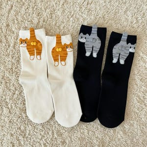 May include: Four pairs of socks with cat designs. Two pairs are white with orange tabby cat designs, and two pairs are black with gray striped cat designs. The cat designs feature the cat's rear end and tail.