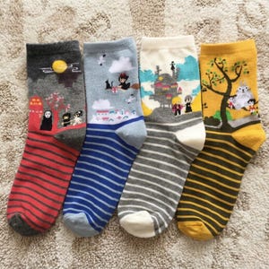 Può includere: Quattro paia di calzini con illustrazioni in stile anime. Ogni calzino presenta un design diverso, inclusi personaggi e scene. I calzini hanno motivi a righe in rosso, blu, grigio e giallo, con vari disegni di personaggi.