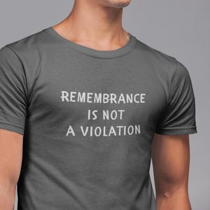 May include: Dark grey t-shirt with the white text "REMEMBERANCE IS NOT A VIOLATION". The shirt has short sleeves and a crew neck. The image is a close-up of the shirt.