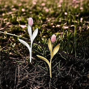 Puede incluir: Dos delicados broches de flor de tulipán, uno en plata y otro en oro, con cabezas de flores rosas. Los broches se muestran sobre un lecho de hierba verde y tierra marrón.