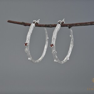 May include: A pair of silver hoop earrings with a textured, branch-like design. Each earring features a small, round, red gemstone. The earrings are displayed against a muted gray background, creating a natural and elegant aesthetic.