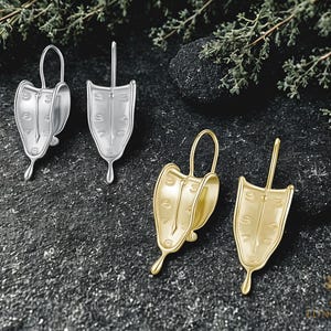 May include: Four earrings in the image. Two are silver, and two are gold. Each earring is shaped like a melting clock, with numbers and a clock hand. The earrings have a unique, surreal design.