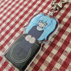 May include: A blue and black glitter keychain featuring a cartoon character with blue hair and a sad expression. The character is wearing a black and white outfit and is sitting down. The keychain is attached to a silver star-shaped charm.