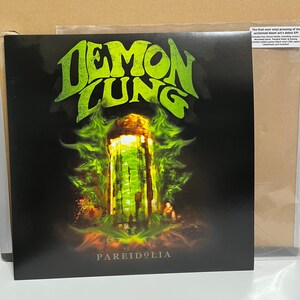 May include: A vinyl record with the text "DEMON LUNG" in green, stylized lettering. The album cover features a green and yellow illustration of a structure with the word "PAREIDOLIA" below. The record is in a protective sleeve.
