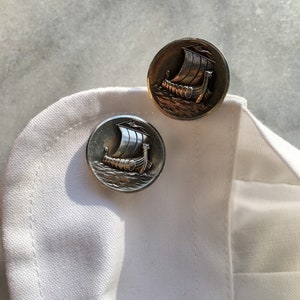 May include: Two silver cufflinks with a raised design of a Viking ship with a sail. The cufflinks are on a white fabric background.