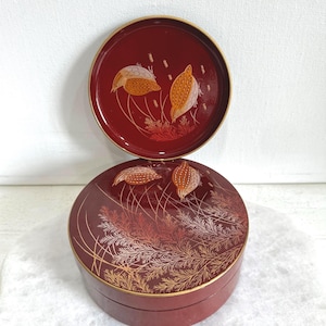 May include: A two-piece, round, burgundy-colored decorative box and lid set. The lid and the top of the box feature a gold-trimmed design with stylized wheat and floral patterns in gold and white.