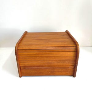 May include: A rectangular wooden bread box featuring a warm, medium-brown wood grain finish. The container is constructed with a horizontal slatted roll-top lid that curves at the sides, providing a smooth, enclosed storage space for kitchen items.