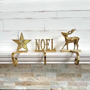 May include: Three brass stocking holders featuring a five-pointed star, the word NOEL in capital letters, and a standing reindeer. Each piece has a curved hook extending downward to hang stockings from a fireplace mantel ledge.