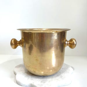 May include: A polished brass ice bucket with a wide, flared rim and two rounded, knob-style handles on opposite sides. The vessel features a smooth, metallic finish with a warm golden hue and a deep, cylindrical body shape.