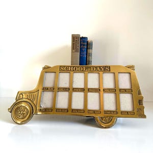 May include: A gold-colored metal school bus-shaped photo frame with spaces for twelve photos, labeled "SCHOOL DAYS" across the top. Each photo slot is labeled with a grade level from 1st to 12th year. Three books are stacked on top of the frame.