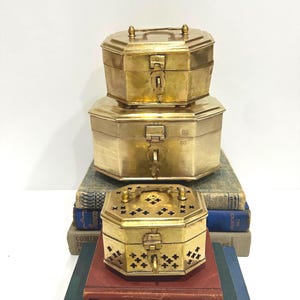 May include: Three antique brass boxes stacked on top of each other. The boxes are octagonal with hinged lids and clasps. The top box has a handle. The bottom box has decorative cutouts. The boxes are sitting on top of old books.