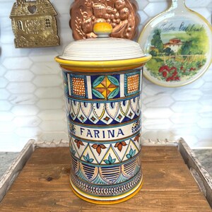 May include: A cylindrical ceramic jar with a lid, featuring a colorful, intricate design. The jar is labeled "FARINA" in a decorative font. The design includes geometric patterns, floral motifs, and a yellow rim. The jar sits on a wooden tray.