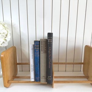May include: A wooden bookend with a light finish holds several vintage books. The books have various colors and titles, including "A Winter Dream" and "A Bright Ray of Darkness". A vase of white flowers sits to the left.