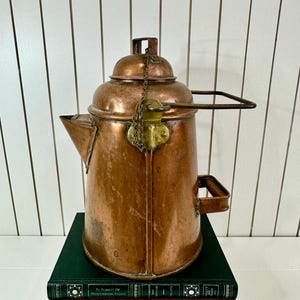 May include: A vintage copper kettle with a brass handle and lid. The kettle is sitting on a stack of books with the titles "Call It Home" and "Live Beautiful".