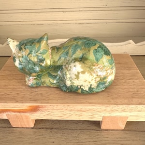 May include: A decorative cat figurine with a green and white leaf pattern, resting on a wooden display stand. The cat is in a relaxed pose, and the stand has a natural wood finish.