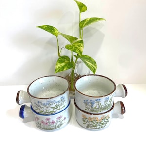 May include: A white ceramic mug with a black handle and the text "But First, Coffee" in a black, handwritten-style font. The mug is on a white surface.