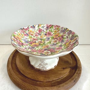 May include: A floral patterned cake stand with a scalloped edge. The stand features a white base with gold accents and a round wooden serving board. The bowl is filled with pink, yellow, and purple flowers.