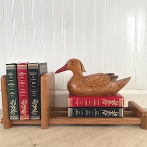 May include: Wooden duck bookends with a carved duck on top of two books. Three books stand between the bookends. The books have red, green, and black covers. The duck has a red beak and is made of light brown wood.