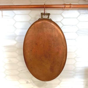 May include: An oval copper pan with a weathered, metallic finish and a single brass handle at the top. The pan is suspended from a horizontal copper rod, showcasing a smooth, rounded surface with natural patina and subtle surface variations.