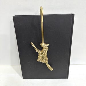 May include: A gold-colored metal witch on a broomstick book holder. The witch is holding a wand and is attached to a long, curved hook. The book holder is attached to a black rectangular object.