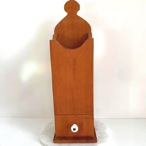May include: A tall, vertical wooden wall-mounted storage bin with a natural light brown wood grain finish. The unit features a curved top cutout, a scalloped front opening, and a small square drawer at the base with a single white ceramic knob.