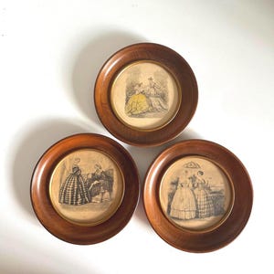May include: Three antique framed prints featuring illustrations of women in period clothing. The round wooden frames have a dark brown finish. The prints depict scenes with women in dresses, some with parasols.
