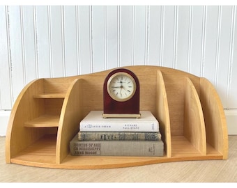 May include: A wooden desk organizer with multiple compartments and shelves. A small, burgundy-colored clock with a gold-colored frame sits atop a stack of books. The organizer is made of light-colored wood and has a curved design.