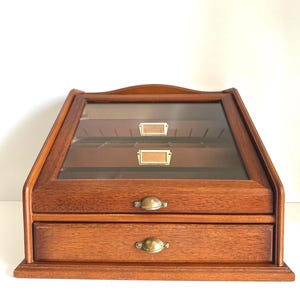 May include: A wooden storage chest with a slanted glass top lid and one lower drawer. The chest features a warm brown wood grain finish, brass-toned metal drawer pulls, and two small rectangular brass label holders on the interior back panel.