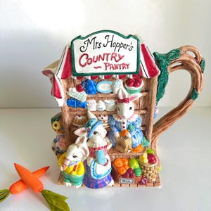 May include: A ceramic teapot shaped like a country pantry, featuring bunny figures and a sign that reads "Mrs. Hopper's Country Pantry." The teapot has a brown handle and is decorated with colorful fruits, vegetables, and baked goods. Two carrot decorations are in the foreground.
