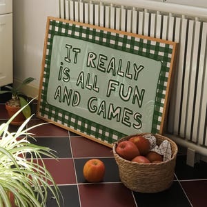 Puede incluir: Un letrero enmarcado con el texto "IT REALLY IS ALL FUN AND GAMES" en una habitación con plantas y una cesta de manzanas. El letrero tiene un borde verde a cuadros y un fondo verde claro.