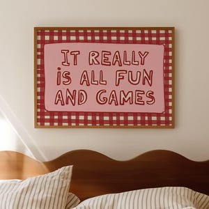 May include: A framed wall art piece with the text "IT REALLY IS ALL FUN AND GAMES" in a playful font. The artwork features a pink background with a red and white checkered border. The art hangs above a bed with striped bedding.