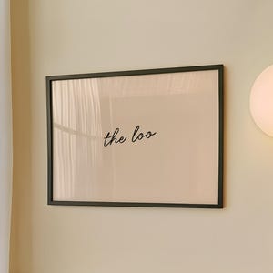 May include: A bathroom scene featuring a framed print with the text "the loo" in cursive. The frame is black, and the print is mounted on a light beige wall. A round, illuminated wall sconce and a light blue tiled wall are also visible.