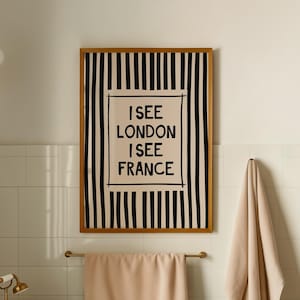 May include: A rectangular framed wall art piece featuring a vertical black and white striped pattern with a central cream-coloured box containing the black text I SEE LONDON I SEE FRANCE. The artwork is displayed on a white tiled bathroom wall.