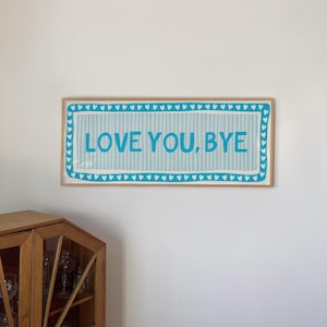 May include: A rectangular wall art piece in a light wood frame featuring a light blue and white vertical striped background. The text LOVE YOU, BYE is printed in bold blue letters, surrounded by a border of small blue hearts on a white background.