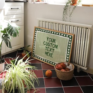 Pu&ograve; includere: Un cartello incorniciato con la scritta "YOUR CUSTOM TEXT HERE" in verde su sfondo a righe, incorniciato da un bordo di cuori verdi. Un cesto di mele e una singola mela si trovano su un pavimento piastrellato rosso e nero.