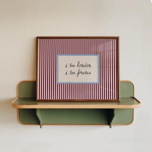 May include: A framed artwork with a brown border, featuring a striped background in red and white. The artwork includes a smaller rectangle with the text "i see london i see france" in cursive. The artwork is displayed on a green shelf.