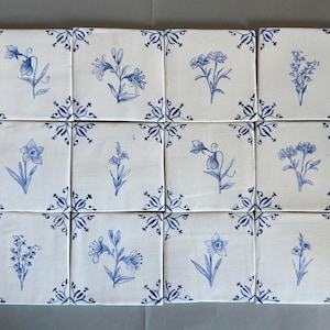 May include: A set of twelve square ceramic tiles arranged in a grid. Each white tile features a unique blue botanical illustration of a flower in the center and blue decorative corner accents. The tiles display various floral designs in blue ink.