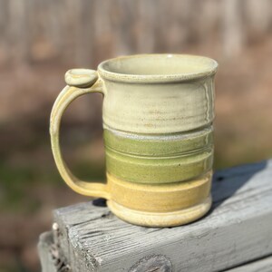 Puede incluir: Una taza de cerámica hecha a mano con un diseño único. La taza presenta bandas horizontales de color crema, verde y amarillo. El asa tiene un reposapulgar distintivo. La taza está sobre una superficie de madera envejecida.