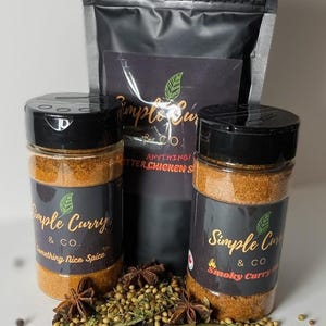May include: A selection of Simple Curry & Co. spice products. Two jars of spice with black lids and labels, a black bag, and a card with text. Spices and herbs are scattered around the jars.