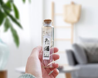 May include: A clear glass bottle with a cork top containing dried flowers and small black and white photos. The bottle is being held by a hand.