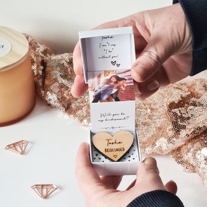 May include: A white box with a heart-shaped wooden piece inside. The wooden piece is engraved with the text "Tasha Bridesmaid". The box is open and the top part of the box has a photo of two women and the text "Jasha I can't say 'I do' without you!" and "Will you be my bridesmaid?"