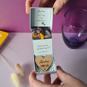 May include: A white box with a heart-shaped wooden cutout that says "Amelia Flower Girl" inside. The box is open and has a photo of a girl and a woman inside. The text on the box reads "Amelia I can't say 'I do' without you!" and "Will you be my flower girl?"