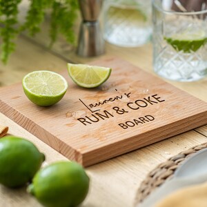 May include: A wooden cutting board with the text "Lauren's Rum & Coke Board" engraved on it. There are two lime wedges and a glass of water on the board.