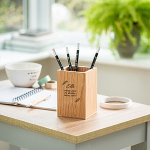 May include: A wooden pencil holder with the name "Ellie" and the quote "Find joy in every new day" engraved on the front. The holder is filled with black pencils.