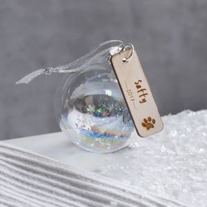 May include: A clear glass bauble with a silver ribbon and a wooden tag that says "Saffy 2017" with a paw print. The bauble is filled with glitter and snowflakes.