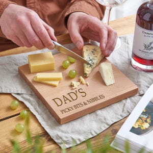 May include: A wooden cutting board with the text "DAD'S PICKY BITS & NIBBLES" engraved on it. The board holds various cheeses, green grapes, and a knife. A bottle of gin-based liqueur is in the background.