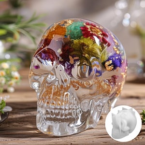 May include: A clear resin skull sculpture with colorful dried flowers and gold leaf inclusions. The skull is transparent, allowing the internal floral arrangement to be visible. The sculpture is a decorative item.